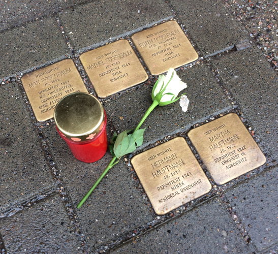 Foto von 5 Stolpersteinen, an denen eine Kerze und eine weiße Rose niedergelegt wurden.