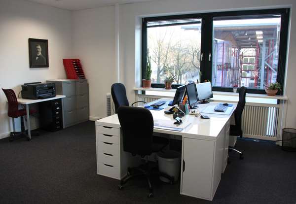 Foto eines Büroraums mit 2 Fenstern, davor ein großer Schreibtisch mit 3 Arbeitsplätzen, links Aktenschränke und ein Drucker.
